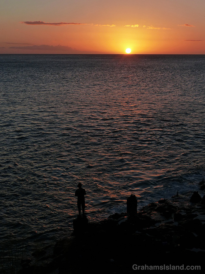 A fishermA fisherman stands on the shore at sunset in Hawaiian stands on the shore at sunset off Hawaii